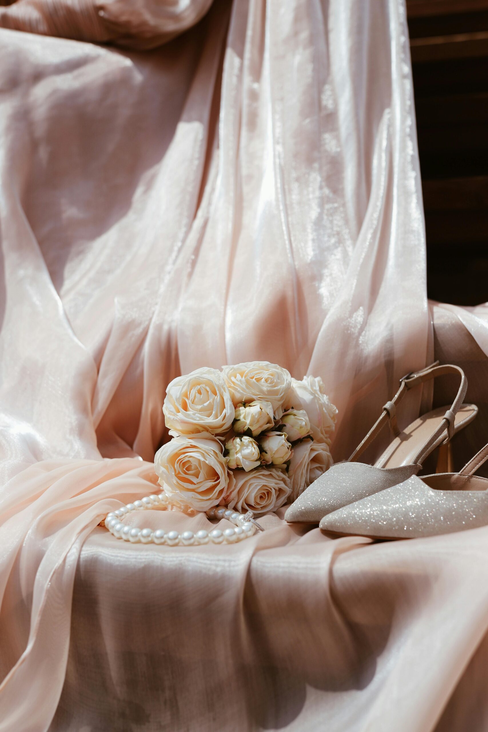 Hochzeitsschuhe, Perlenkette und Brautstrauß aus weißen Rosen auf edlem Stoff drapiert